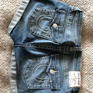 Blå jeansshorts True Religion - Säljer ett par blå jeansshorts från True Religion med klassiska kontrastsömmar och stora bakfickor med lock och knapp. Shortsen har upprullade benslut och en snygg, avslappnad passform. Perfekta för hösten med strumpbyxor under och riktigt ikoniska detaljer.🙏