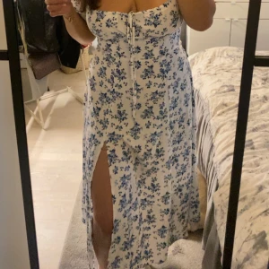 Vit långklänning med blå blommor - Supersöt långklänning i vitt med blått blommönster. Klänningen har snörning vid bysten, korta puffärmar och en snygg slits framtill och dragkedja på ryggen. Perfekt för sommarens alla tillfällen och ger en romantisk vibe.