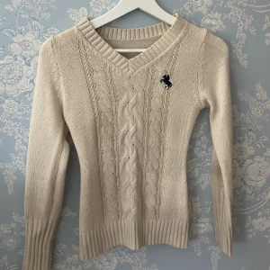 Beige stickad tröja  - Snygg beige stickad tröja  med kabelstickat mönster och marinblå broderad logga på bröstet. Tröjan har v-ringning, långa ärmar och breda ribbade muddar vid ärmslut och nederkant. Perfekt för lager-på-lager under kyliga dagar. Slim .