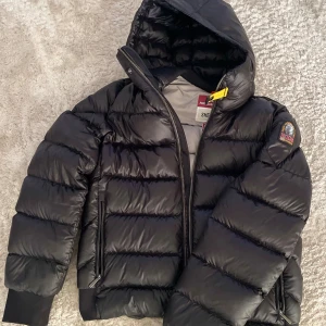 Svart pufferjacka Parajumpers M - Svart pufferjacka från Parajumpers, modell Sheen, med huva och snygga detaljer som gul rem vid halsen och patch på ärmen. Jackan har dragkedja, två sidofickor med zip och ribbade muddar. Perfekt inför den svenska vintern som börjar närma sig och speciellt för dig som vill hålla en stilren look. Ny pris för jackan är ca 7000kr. För fler bilder på självaste jackan är det bara att kontakta mig!