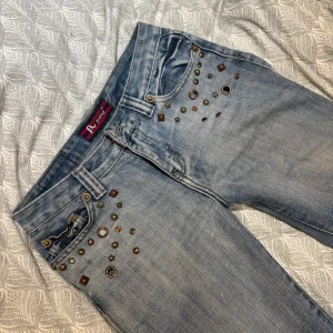 Ljusblå bootcut jeans med nitar - Säljer ett par ljusblå jeans från A ping med bootcut-modell och låg midja. Jeansen har coola nitar och metalldetaljer i olika former och storlekar framtill vid fickorna. Tillverkade i klassiskt denimtyg och perfekta för dig som vill sticka ut. De är ascoola men tyvärr lite för korta enligt mig, de går typ precis ner till marken för mig som är 166. De har små rostfläckar längst ner som kommit från regnet, och de är uppsydda men inte klippta där nere. Midjemått: 38/39, innerben: 72