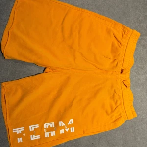 Gula shorts från H&M med TEAM-tryck - Snygga gula shorts från H&M med vitt TEAM VIBES-tryck på ena benet. De har elastisk midja och är gjorda i mjuk bomull, perfekta för chill dagar eller sport. Klassisk passform och skön känsla hela sommaren.