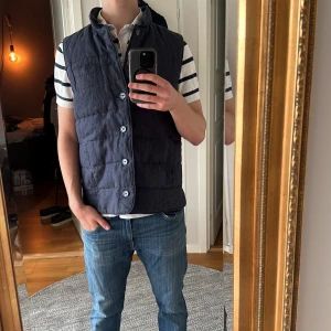 John Henric mörkblå dunvänst - Snygg mörkblå quiltad dunväst från John Henric med ståkrage och knappar framtill. Västen har ett stilrent, rakt snitt och är tillverkad i linne med polyesterfoder och fyllning. Perfekt att slänga över en t-shirt eller hoodie för extra värme.