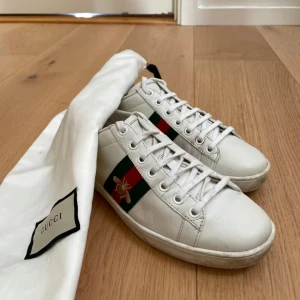 Gucci Ace vita sneakers med bi - Gucci Ace sneakers i vitt skinn med klassiska gröna och röda ränder på sidan samt broderad guldfärgad bi. Baksidan har metallicröd och metallicgrön detalj med Gucci-logga. Platt sula och snörning framtill. Skit snygga men används inte längre, kvitto kommer med💞