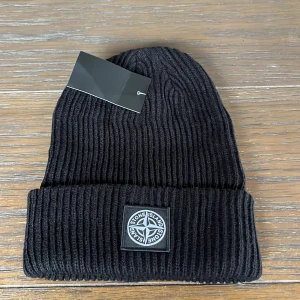 Svart ribbad mössa från Stone Island - Svart ribbstickad mössa från Stone Island med klassisk logopatch framtill. Snygg och enkel design som passar till allt, perfekt för kalla dagar. Materialet är stickat och mössan har en uppvikt kant för extra stilpoäng.