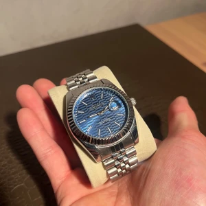 R-klocka  blå randig urtavla - R-Oyster Perpetual Datejust med en snygg blå urtavla som har ett randigt mönster. Klockan har en klassisk rund form, räfflad bezel och länkarmband i silverfärgat stål. Datumvisning med förstoringsglas och tydliga indexmarkeringar. Perfekt för dig som gillar stil och kvalitet.