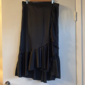 Svart omlottkjol med volang från MAPP - Svart lång omlottkjol från MAPP jeans i glansigt, mjukt material. Kjolen har volangdetaljer längs nederkanten och knyts i midjan för en snygg passform. Perfekt för dig som gillar en flowy och elegant look.