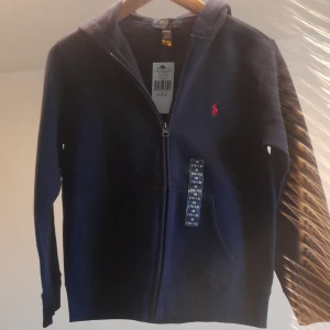 Marinblå hoodie från Polo Ralph Lauren - Snygg marinblå hoodie från Polo Ralph Lauren med dragkedja och huva. Klassisk design med röd broderad logga på bröstet och känguruficka framtill. Perfekt för en chill och stilren look.