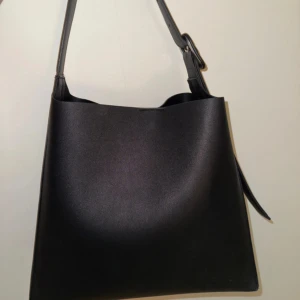 Svart väska - Stilren svart väska  med justerbart axelband och magnetlås. Väskan har en rymlig insida och ett innerfack med dragkedja för extra säkerhet. Perfekt för dig som gillar minimalistisk och modern design. Perfekt nu till hösten🍁