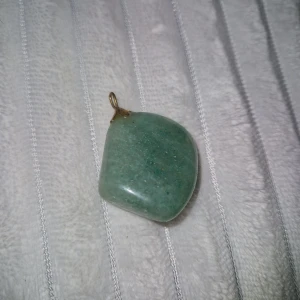 Kristall/sten  - Unikt hänge i grön sten/kristall med mjukt rundad form och ett litet fäste i guldfärgad metall. Perfekt att sätta på en kedja och bära som halsband. Stenen har en naturlig, lite skimrande yta som ger en cool vibe.