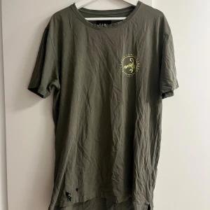 Olivgrön t-shirt från CSBL XL - Olivgrön t-shirt från CSBL i storlek XL med tryck i gult framtill och stort statement-tryck på ryggen. Klassisk rund hals och korta ärmar. Skön och avslappnad passform, perfekt för en streetwear-look.