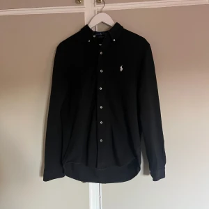 Svart skjorta från Ralph Lauren M - Svart långärmad skjorta från Ralph Lauren i modellen Featherweight Mesh. Skjortan har klassisk krage, knäppning framtill med vita knappar och den ikoniska broderade loggan på bröstet. Tillverkad i ett lätt och luftigt bomullsmaterial.