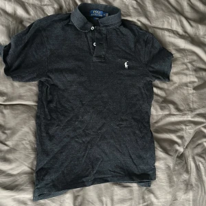 Svart pikétröja Polo Ralph Lauren M - Svart/grå pikétröja från Polo Ralph Lauren i slim fit med klassisk krage och knappar framtill. Tröjan har korta ärmar och den ikoniska broderade loggan på bröstet. Perfekt för en clean och stilren look. Den är väldigt liten i storlek så passar bättre som S