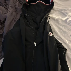 Svart vindjacka från Moncler - Svart vindjacka från Moncler med klassisk logga på ärmen och snygga rödvita detaljer i huvan. Jackan har dragkedja framtill och justerbar huva. Perfekt för dig som vill ha en stilren och sportig look.