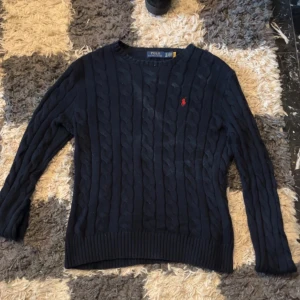 Mörkblå kabelstickad tröja Polo Ralph Lauren - Kabelstickad mörkblå tröja från Polo Ralph Lauren med klassisk rund halsringning och röd broderad logga på bröstet. Tröjan har långa ärmar och ribbade muddar vid ärmslut och nederkant. Perfekt för en stilren och tidlös look.