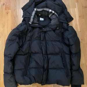 Svart pufferjacka från Burberry M - Säljer en svart Burberry dunjacka i storlek M. Jackan är använd väldigt få gånger och är i toppskick. Den har en klassisk pufferdesign med huva, dragkedja och knappar framtill samt två praktiska fickor med dragkedja. Insidan av kragen har det ikoniska grå rutiga fodret som ger en snygg och exklusiv kontrast. 