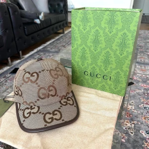 Gucci beige keps med GG-mönster - Snygg beige keps från Gucci med brunt GG-logomönster över hela kepsen. Kepsen har en böjd skärm med brunt kantband och justerbar rem baktill i mörkbrunt skinn. Materialet är slitstark canvas och detaljerna ger en lyxig känsla. Perfekt för dig som gillar designeraccessoarer.