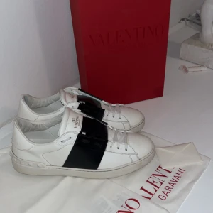 Valentino Open sneakers vit/svart - Snygga Valentino Open sneakers i vitt skinn med bred svart läderdetalj över mitten. Klassisk rund tå, platt sula och snörning framtill. Perfekt för dig som gillar stilrena och lyxiga sneakers med en unik touch. Kan även sätta på extra snören och tvätta dom så att dom ser så gott som nya ut.
