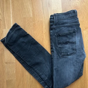 Svarta Nudie Jeans Grim Tim 30/32 - Snygga svarta Nudie Jeans Grim Tim i tvättad look. Klassisk femficksmodell med raka ben och normal passform. Jeansen är tillverkade i stretchig bomullsmix för extra komfort och har diskreta slitningar för en cool vibe.