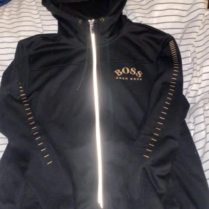 Svart vindjacka från Hugo Boss L - Svart vindjacka från Hugo Boss med guldiga detaljer och logga på bröstet. Jackan har huva, hel dragkedja framtill och dekorativa guldstreck längs ärmarna. Tillverkad i polyester för en sportig och clean look. Perfekt för dig som gillar stilrena streetwear-plagg.