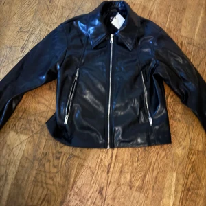 Svart skinnjacka från Zara, S - Snygg svart skinnjacka från Zara i storlek S. Jackan har klassisk krage, raka dragkedjor framtill och en stilren, croppad passform. Perfekt för dig som vill ha en edgy look med stilrena detaljer och glansig finish.