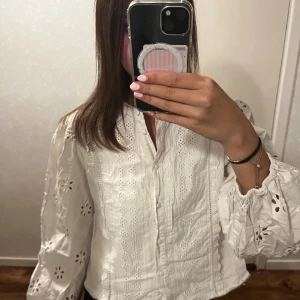 Vit broderad blus från Zara - Superfin vit blus från Zara med broderade detaljer och hålmönster. Blusen har långa puffärmar med resår vid ärmslut och knappar framtill. Perfekt för dig som gillar romantisk och trendig stil.