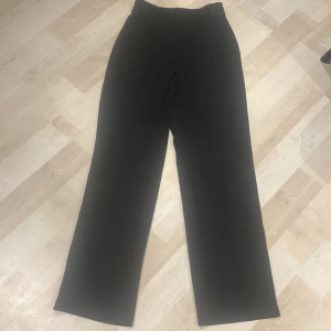 Svarta vida kostymbyxor - Stilrena svarta kostymbyxor med hög midja och vida ben. Klassisk look med bälteshällor och pressveck framtill. Perfekta för en clean och snygg outfit. Materialet känns mjukt och följsamt.