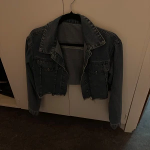 Croppad jeansjacka i blå denim - Säljer en croppad jeansjacka i klassisk blå denim med rå kant nertill. Jackan har två bröstfickor med knappar, bred krage och långa ärmar. Perfekt för dig som vill ha en edgy och trendig look.
