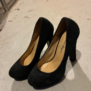 Svarta pumps från Claudia Ghizzani - Klassiska svarta pumps från Claudia Ghizzani med rund tå och hög klack. Skorna är i mocka och har en stilren, tidlös design som passar till många olika outfits. Perfekta för dig som gillar en elegant look med lite extra höjd.