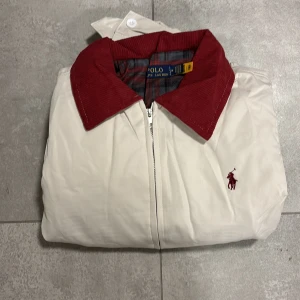 Vit vindjacka från Polo Ralph Lauren - Vit vindjacka från Polo Ralph Lauren med röd manchesterkrage och broderad logga på bröstet. Jackan har dragkedja framtill, två fickor och elastisk midja. Perfekt för dig som vill ha en stilren och klassisk look med en twist. Passar mer som S än M enligt mig.