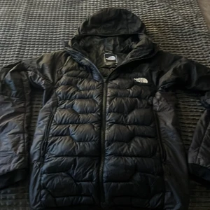 Svart pufferjacka från The North Face - Helt ny Svart pufferjacka från The North Face med huva och dragkedja. Jackan har quiltad design och är tillverkad i syntetmaterial, perfekt för kalla dagar. Klassisk logga på bröstet och snygg passform som funkar till streetwear.