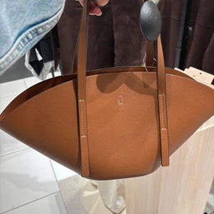 Brun handväska i skinn - Snygg brun handväska i skinn med stilren och modern form. Väskan har långa handtag och en minimalistisk design utan synliga loggor. Perfekt för dig som gillar en clean look och vill ha plats för allt du behöver under dagen.