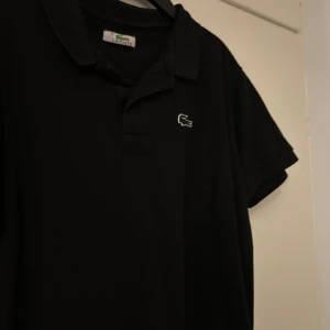 Svart pikétröja från Lacoste - Klassisk svart pikétröja från Lacoste med ikoniska krokodilloggan broderad på bröstet. Tröjan har krage, knäppning framtill och korta ärmar. Perfekt för en clean och stilren look. Materialet är mjukt och andas, vilket gör den skön att bära. Priset kan diskuteras 