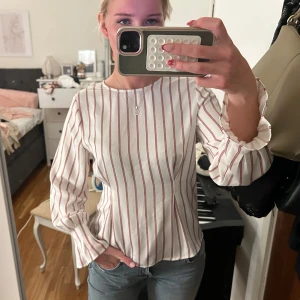 Randig blus från Gina Tricot - Vit blus med röda vertikala ränder från Gina Tricot. Modellen har långa ärmar med volangdetalj vid ärmslut och en rak passform. Tunt, lätt material som känns luftigt och fräscht. Perfekt att styla med jeans eller kjol för en trendig look.