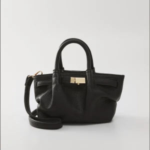 Svart handväska med axelrem - Handväska från Zalando, lik den bruna populära från Lindex. Snygg handväska i svart och brun med justerbar axelrem och guldfärgade metalldetaljer. Väskan har två handtag, struktur i skinn och praktisk öppning med dekorativ låsdetalj. Perfekt storlek för det viktigaste och stilren design som lyfter din outfit.