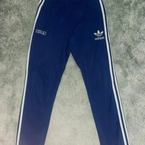 Blå Adidas Italien byxor - Snygga blå byxor från Adidas med vita ränder längs sidorna och broderad Adidas-logga samt 'Italia' på ena benet. Byxorna har elastisk midja och är tillverkade i ett glansigt syntetmaterial. Perfekta för sköna outfits. Kan gå ner i pris. Sällsynta❗️