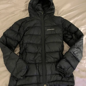 Svart dunjacka Peak Performance XS - Svart dunjacka från Peak Performance i storlek XS. Jackan har huva, quiltad design och dragkedja framtill. Perfekt för kalla dagar med sin lätta och puffiga stil. Snygg logga på bröstet och långärmad modell. Den har haft ett hål på insidan av armen men som jag lagat med en liten svart lapp, så det syns knappt 🤗