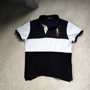 Randig pikétröja från Polo Ralph Lauren - Snygg pikétröja från Polo Ralph Lauren i svart och vitt med breda ränder. Klassisk krage, korta ärmar och stor broderad polospelare på bröstet. 