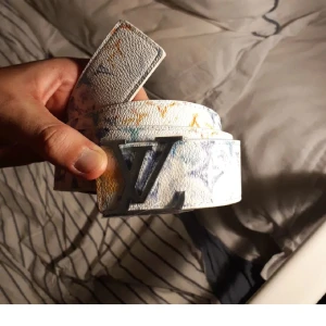 Louis Vuitton vitt bälte med färgstänk - Säljer ett Louis Vuitton bälte i vitt skinn med coola färgstänk i blått, gult och orange. Bältet har en stor LV-logga i silver som spänne och insidan är ljusbrun. Perfekt accessoar för att lyfta din outfit.