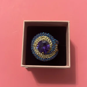 Statementring med lila sten och blå detaljer - Cool statementring med stor lila sten i mitten, omgiven av glittriga blå och guldiga stenar. Ringen har en rund form och är riktigt iögonfallande med sin färgkombination och bling. Perfekt för dig som vill sticka ut och addera färg till din look.