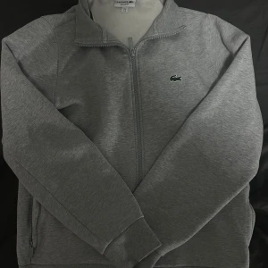 Lacoste tröja grå  - Snygg grå ziptröja från Lacoste med klassisk krokodillogga på bröstet. Tröjan har hel dragkedja, ståkrage och är fodrad med mjuk fleece på insidan. Perfekt för kyliga dagar och enkel att matcha med det mesta.