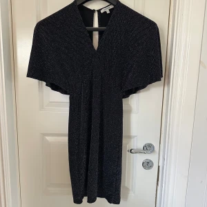 Glittrig svart kortklänning Chiquelle XS - Svart kortklänning från Chiquelle i storlek XS med glittriga silverdetaljer. Klänningen har v-ringning, vida korta ärmar och en snygg öppen rygg med knapp i nacken. Perfekt för dig som vill sticka ut med en elegant och trendig look. 