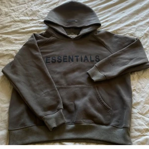 Brun Essentials Fear of God hoodie - Säljer en brun hoodie från Essentials Fear of God med svart logga på bröstet och ärmen. Tröjan har huva, känguruficka och är tillverkad i mjuk bomullsmix. Perfekt för dig som gillar streetwear och vill ha en clean look.