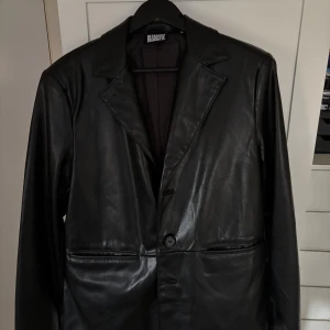 Svart skinnkavaj Reclaimed Vintage M - Svart kavaj i konst-skinn från Reclaimed Vintage, storlek M. Klassisk design med breda slag, två framfickor och en knappstängning. Snygg och stilren look med slät yta och diskreta detaljer. Perfekt för dig som vill ha en edgy och cool vibe.