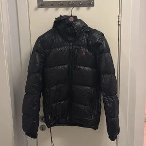 Mörkblå pufferjacka Polo Ralph Lauren - Mörkblå pufferjacka från Polo Ralph Lauren med röd broderad logga på bröstet. Jackan har huva, dragkedjor framtill och på fickor samt quiltad design. Perfekt för kalla dagar och har en sportig, clean look. Priset kan diskuteras vid snabb affär.