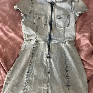 Kort jeansklänning med dragkedja Lindex - Snygg ljusblå jeansklänning från Lindex med korta ärmar, ståkrage och två bröstfickor. Dragkedja framtill med ringdetalj och markerade sömmar ger en cool vibe. Perfekt för dig som gillar denim och vill ha en unik look.
