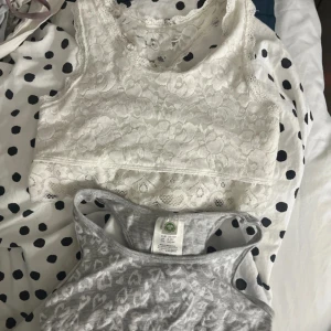 Vit spetsig linnetopp & grå hjärtmönstrad topp - Två toppar från Lindex: en vit linnetopp i spets med blommigt mönster och racerback, samt en grå linnetopp med vita hjärtan och sportig rygg. Båda är ärmlösa och har en croppad passform, perfekta för att ge en chill vibe till din outfit.