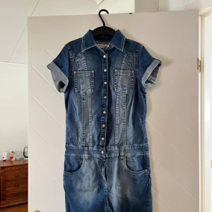 Jeansklänning från Denim 36 med knappar - Snygg jeansklänning från Denim 36 med korta ärmar, knappar hela vägen framtill och två bröstfickor. Klassisk skjortkrage och coola sömdetaljer. Perfekt för dig som gillar denim och vill ha en avslappnad men trendig look.