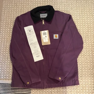 Lila Carhartt Jacka - Lila Carhartt jacka som är helt oanvänd. Köpt på Naturkompaniet i våras men glömde helt att använda den så för helst tillbaka en del av pengarna så man kan droppa på lite annat. Riktigt skönt starkt material med skön insida. Carhartts klassiska tjocka dragkedja och guldknappar. Sitter riktigt bra på modellen som är 179 65kg. Skickar eller möts i Stockholm. 