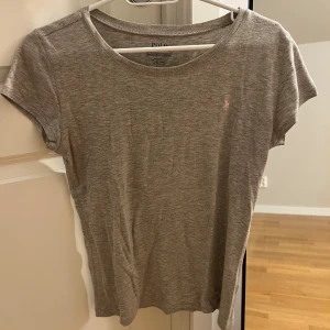 Grå t-shirt från Polo Ralph Lauren - Snygg grå t-shirt från Polo Ralph Lauren med liten rosa logga broderad på bröstet. T-shirten har rund halsringning och korta ärmar. Perfekt basic-plagg i mjuk bomull med normal passform. Tröjan är i barnstoleken men passar som storlek s. 